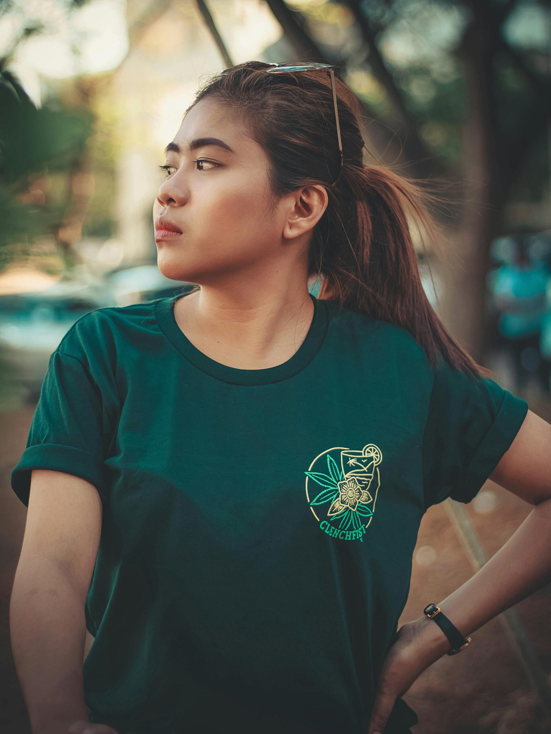 A woman wearing a green T-shirt stands outdoors, gazing thoughtfully to the side.